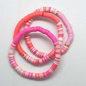 Op de afbeelding: Drie gestapelde armbanden gemaakt van roze, witte en oranje kralen. Elke armband heeft een gouden accent.