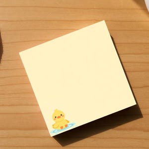 May include: A yellow sticky note with a small cartoon duck illustration in the corner. The note is on a wooden surface, next to a white ceramic cup and a white cloth. The duck is yellow with an orange beak and is in blue water.