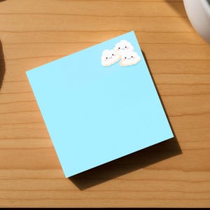 May include: A light blue sticky note with two small cloud-shaped stickers on the top left corner. The note is on a wooden cutting board, with a white cup and a white cloth in the background.