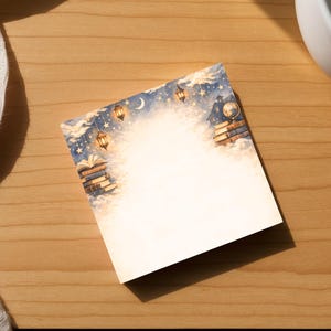 May include: A square, blank card featuring a watercolour illustration of books, lanterns, and a night sky. The card is on a wooden surface, beside a white ceramic mug and a white cloth.