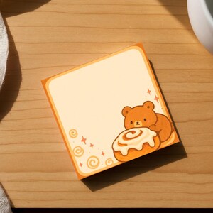 May include: A square sticky note with an orange border and a cartoon bear hugging a cinnamon roll. The note is on a wooden surface, next to a white mug and a white cloth.