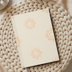 May include: A cream-colored journal with a sunburst pattern in orange ink rests on a woven beige mat. A cup of coffee and dried flowers are also visible, creating a warm, inviting aesthetic. The journal has a simple, minimalist design.