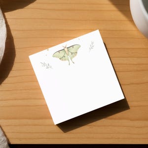 May include: A square white notepad with a watercolor illustration of a green moth and sprigs. The notepad is on a wooden surface, next to a white mug and a white cloth.