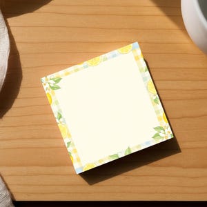 May include: A square notepad with a lemon and leaf border rests on a wooden surface. The notepad has a blank cream-colored center. A white ceramic cup and a white cloth are also in the image.