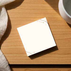 May include: A square, off-white card with delicate gold-colored line art and small dots in the corners rests on a wooden surface. A white ceramic cup and a draped, off-white fabric are also present, creating a minimalist aesthetic.