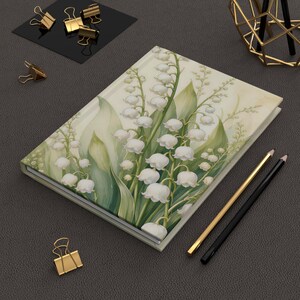 May include: A rectangular notebook featuring a botanical illustration of white lily of the valley flowers with green leaves on a cream background. The notebook is positioned alongside gold-toned metal binder clips, a gold pen, a black pen, and a gold wire holder.