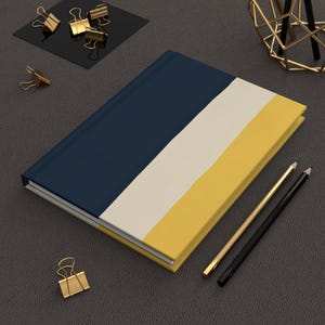 May include: A hardcover notebook featuring a tri-color design with diagonal stripes in navy blue, cream, and mustard yellow. The notebook is accompanied by one gold-toned pencil and one black pencil, along with several gold-toned metal binder clips.