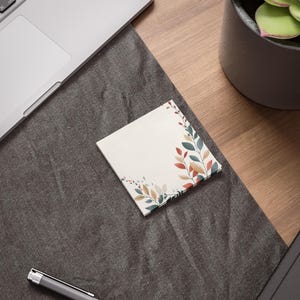 May include: A white square sticky note with a floral design featuring red, orange, and green leaves. The note is on a gray surface.
