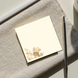 May include: A square, cream-colored sticky note featuring a watercolor illustration in the bottom left corner. The graphic depicts a light brown rabbit wearing a green scarf, pushing a wooden wheelbarrow filled with pink and yellow tulips.