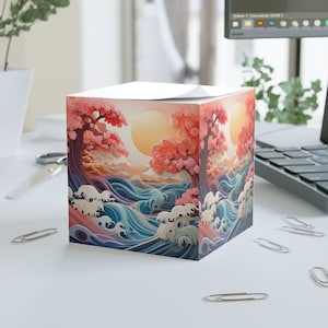 Puede incluir: Un bloc de notas cuadrado con una ilustración colorida de una escena de inspiración japonesa. El diseño presenta un océano estilizado con olas, un sol naciente y dos cerezos en flor. Los colores son vibrantes e incluyen tonos de rosa, azul, verde y naranja.