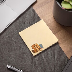May include: A square, light yellow sticky note featuring a cartoon illustration of a brown beaver holding a yellow pencil and a small blue notebook. The beaver is positioned in the bottom corner of the note, which has a smooth, matte surface.