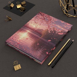 Può includere: Un diario con copertina rigida con una serena scena paesaggistica, un ponte sull'acqua, fiori di ciliegio e un tramonto. Sono visibili anche matite dorate e nere e fermagli dorati.