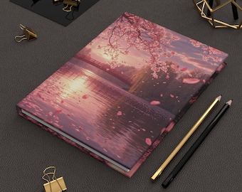 May include: A hardcover journal featuring a serene landscape with a bridge over water, cherry blossoms, and a sunset. The cover is predominantly pink and purple. Gold and black pencils and gold binder clips are also visible.