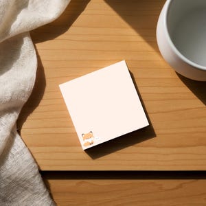 May include: A square, light peach-colored sticky note with a small cartoon fox illustration in the corner. The note is on a wooden surface, next to a white ceramic cup and a white cloth.