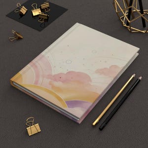 May include: A hardcover notebook featuring a soft-toned illustration of clouds, sun rays, and circular motifs in shades of peach, pink, and purple. The notebook is accompanied by a gold-toned pencil, a black pencil, and several gold-toned metal binder clips.