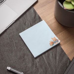 May include: A square light blue sticky note featuring a small illustration of a tan mouse wearing glasses and a scarf, reading a blue book while sitting next to a stack of three books and a steaming cup of coffee in the bottom right corner.