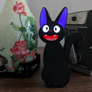 Pode incluir: Uma estatueta preta em forma de gato com orelhas roxas e boca vermelha. O gato est&aacute; em uma superf&iacute;cie de madeira, com um vaso decorativo e uma c&acirc;mera vintage ao fundo. O vaso tem um design floral.