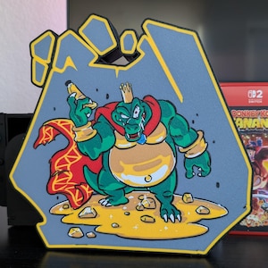 May include: A gray, hand-shaped decorative piece featuring a cartoon image of King K. Rool from the Donkey Kong video game series. The character is green with a gold crown and red cape, holding a banana. A Nintendo Switch game is visible.