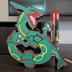 K&ouml;nnte beinhalten: Eine gr&uuml;ne und gelbe Rayquaza-Figur, ein schlangenartiges, drachen&auml;hnliches Pok&eacute;mon, mit roten Akzenten an Maul und Bauch. Die Figur steht auf einer dunklen Oberfl&auml;che, im Hintergrund eine Nintendo Switch-Konsole und Spielh&uuml;llen.