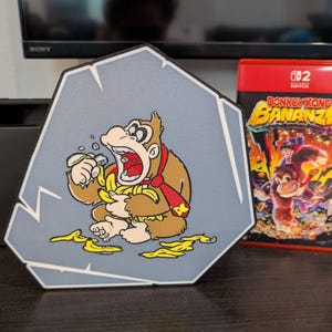 May include: A gray, rock-shaped decorative item featuring a cartoon Donkey Kong character eating bananas. A red Nintendo Switch game case with the title "Donkey Kong Bananza" is next to it.