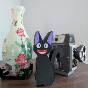 Pode incluir: Uma estatueta de gato preto com orelhas roxas e boca vermelha. Uma c&acirc;mera de filme vintage e um vaso decorativo com desenhos florais e de escrita tamb&eacute;m est&atilde;o presentes.