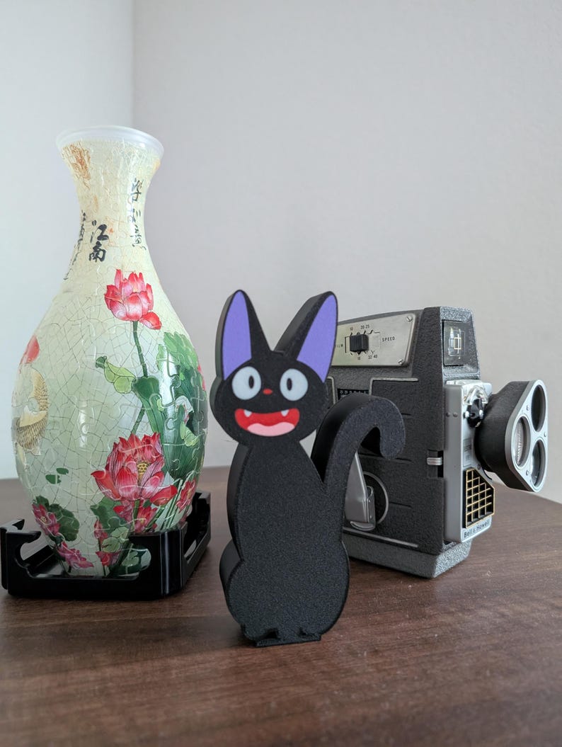 Pode incluir: Uma estatueta de gato preto com orelhas roxas e boca vermelha. Uma c&acirc;mera de filme vintage e um vaso decorativo com desenhos florais e de escrita tamb&eacute;m est&atilde;o presentes.