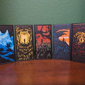 May include: Five framed art prints with fantasy-themed illustrations. Each print features a different design: a wolf, a lion, a stag, a raven, and a forest scene. The color palette includes blue, yellow, red, orange, and black.