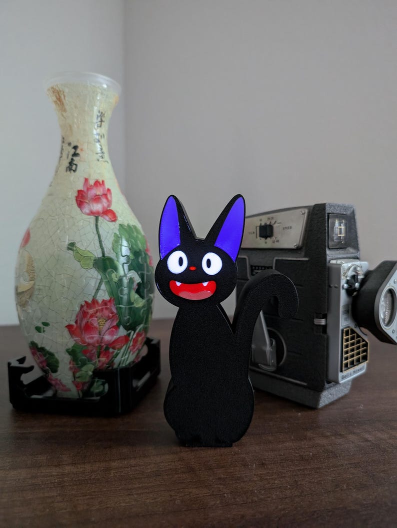 Pode incluir: Uma estatueta preta em forma de gato com orelhas roxas e boca vermelha. O gato est&aacute; em uma superf&iacute;cie de madeira, com um vaso decorativo e uma c&acirc;mera vintage ao fundo. O vaso tem um design floral.