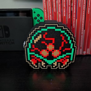 May include: A pixelated black, green, red, and tan accessory case, likely for a Nintendo Switch. The case features a retro video game design. A black Nintendo Switch console and game cartridges are in the background.