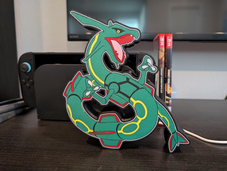 K&ouml;nnte beinhalten: Eine gr&uuml;ne und gelbe Rayquaza-Figur, ein schlangenartiges, drachen&auml;hnliches Pok&eacute;mon, mit roten Akzenten an Maul und Bauch. Die Figur steht auf einer dunklen Oberfl&auml;che, im Hintergrund eine Nintendo Switch-Konsole und Spielh&uuml;llen.