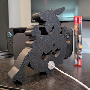K&ouml;nnte beinhalten: Schwarzer, 3D-gedruckter, zeichenf&ouml;rmiger St&auml;nder f&uuml;r ein Telefon oder eine Spielkonsole. Ein wei&szlig;es Ladekabel ist mit dem St&auml;nder verbunden. Im Hintergrund sind zwei Spielkassetten sowie eine graue Spielkonsole zu sehen.