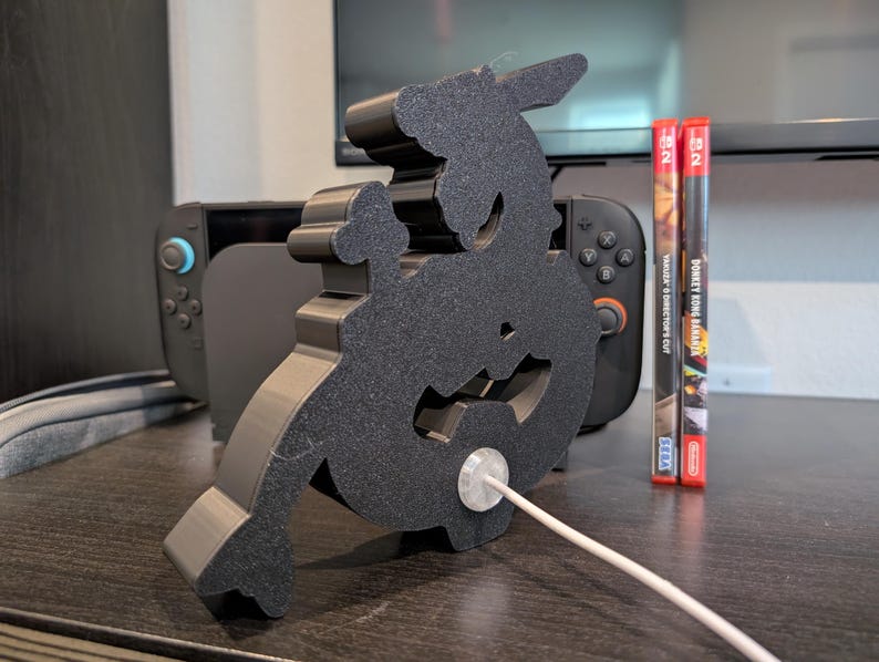 K&ouml;nnte beinhalten: Schwarzer, 3D-gedruckter, zeichenf&ouml;rmiger St&auml;nder f&uuml;r ein Telefon oder eine Spielkonsole. Ein wei&szlig;es Ladekabel ist mit dem St&auml;nder verbunden. Im Hintergrund sind zwei Spielkassetten sowie eine graue Spielkonsole zu sehen.