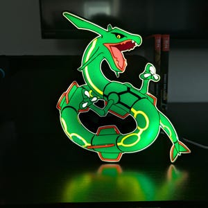 K&ouml;nnte beinhalten: Eine gr&uuml;n und gelb beleuchtete Rayquaza-Figur, eine Figur aus dem Pok&eacute;mon-Franchise. Die Figur hat einen roten Mund und Akzente mit einer leuchtenden Kontur. Die Figur befindet sich auf einer schwarzen Oberfl&auml;che.