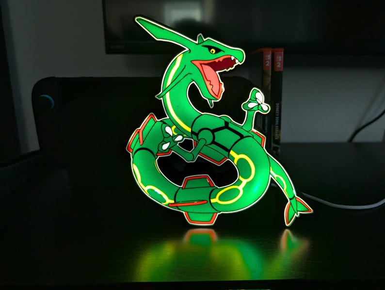 K&ouml;nnte beinhalten: Eine gr&uuml;n und gelb beleuchtete Rayquaza-Figur, eine Figur aus dem Pok&eacute;mon-Franchise. Die Figur hat einen roten Mund und Akzente mit einer leuchtenden Kontur. Die Figur befindet sich auf einer schwarzen Oberfl&auml;che.