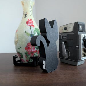 Pode incluir: Um objeto decorativo preto em forma de gato est&aacute; ao lado de uma c&acirc;mera vintage e um vaso floral. O vaso &eacute; creme com desenhos florais vermelhos e rosa. A c&acirc;mera &eacute; cinza e preta.