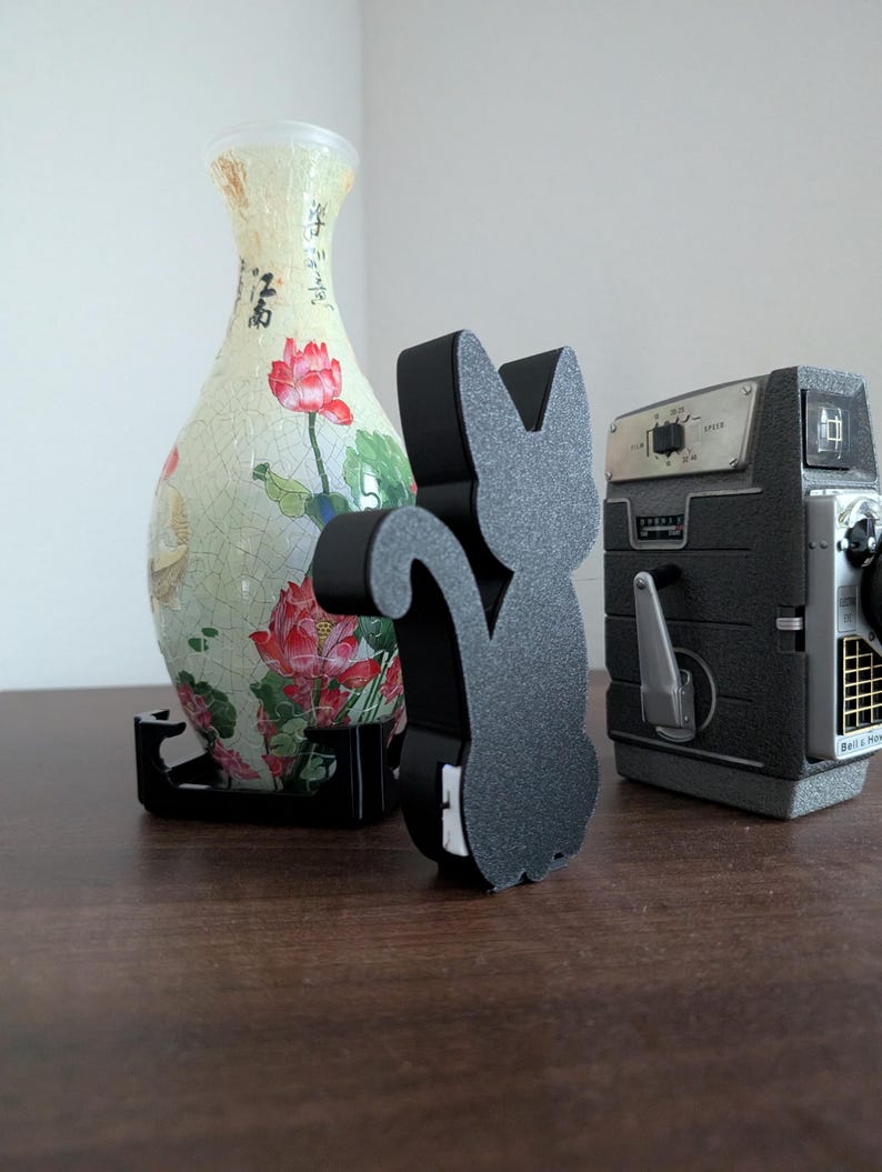 Pode incluir: Um objeto decorativo preto em forma de gato est&aacute; ao lado de uma c&acirc;mera vintage e um vaso floral. O vaso &eacute; creme com desenhos florais vermelhos e rosa. A c&acirc;mera &eacute; cinza e preta.