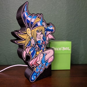 May include: A 3D-printed figure of a female anime character in a blue and pink outfit, holding a staff. The figure is mounted on a black base. A green "Deck Box" is in the background.