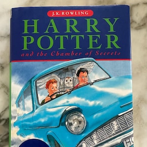 Puede incluir: Una cubierta de libro azul con el título "Harry Potter y la Cámara Secreta" de J.K. Rowling. La cubierta presenta una ilustración de un coche azul volando con un búho blanco y dos niños pequeños dentro.