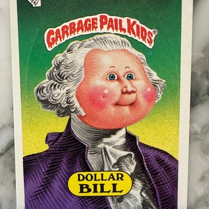 Może przedstawiać: Karta kolekcjonerska Garbage Pail Kids przedstawiająca kreskówkową ilustrację osoby z dużą głową i różowymi policzkami. Karta jest zielona i żółta z tekstem "Garbage Pail Kids" i "Dollar Bill" na niej. Numer karty to 73b.