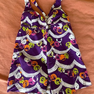May include: A purple swimsuit with a white and colourful abstract print. The swimsuit has a V-neckline and thin straps.