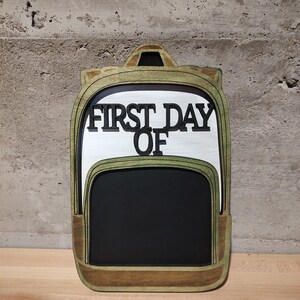 May include: Wooden cutout of a backpack with a chalkboard insert. The backpack is green and brown with the words "FIRST DAY OF" written on the front.