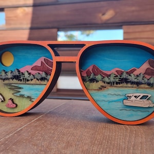May include: A wooden pair of sunglasses with a layered scene of a lake, mountains, and a boat inside the lenses. The sunglasses are orange with a black frame.