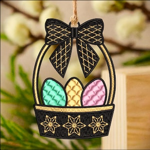 May include: Embroidered Easter basket ornament with a black bow and gold accents. The basket holds three colorful eggs: green, yellow, and pink. The basket has a black base with gold star designs. The ornament hangs from a natural twine.