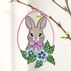 Könnte beinhalten: Ein ovaler rosa Rahmen mit einem gestickten grauen Hasen mit langen Ohren, einer rosa Nase und einer violetten Fliege. Der Hase ist von blauen Blumen mit gelben Mitten und grünen Blättern umgeben, was ein dekoratives Hängeornament ergibt.