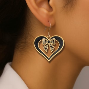 Puede incluir: Pendientes dorados en forma de corazón con interior negro y diseño de lazo. Los pendientes cuelgan de un gancho plateado y se llevan en la oreja. Los pendientes tienen un aspecto detallado y texturizado.