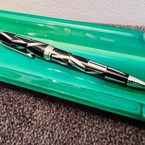 Könnte beinhalten: Ein gemusterter Stift liegt in einer hellgrünen rechteckigen Schale mit Zierkanten. Der Stift hat ein schwarz-silbernes Design. Die Schale ist in einem leuchtenden Grünton gehalten. Der Stift ist aus Metall.