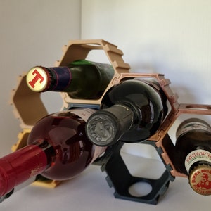 Puede incluir: Un estante hexagonal para vino hecho de plástico marrón y negro contiene tres botellas de vino y una botella de cerveza. Las botellas de vino son roja, blanca y rosada. La botella de cerveza está etiquetada como "Birra Moretti".