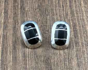 May include: A pair of silver-toned stud earrings with a rectangular shape. Each earring features a black, geometric design with a horizontal silver accent. The earrings are displayed on a wooden surface.