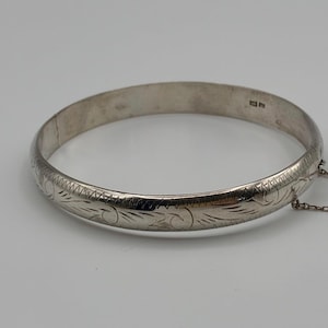 Puede incluir: Una pulsera rígida de plata con un patrón floral grabado en la superficie exterior. La pulsera incluye un mecanismo de apertura con bisagra, una pequeña cadena de seguridad adjunta y sellos distintivos visibles en la banda interior.