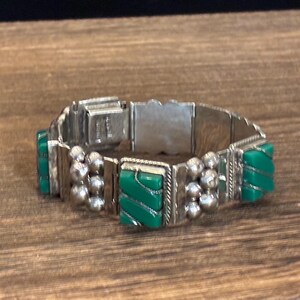 May include: Silver-toned bracelet with rectangular green accents and spherical silver details. The bracelet is made of multiple connected segments and is resting on a wooden surface.